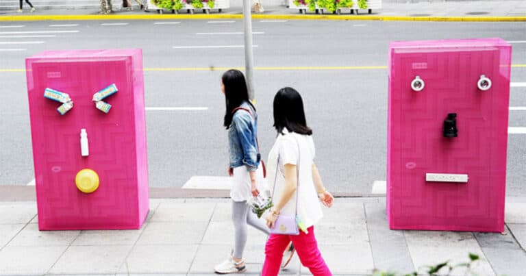 two people walking on sidewalk by large pink boxes that have faces on them
